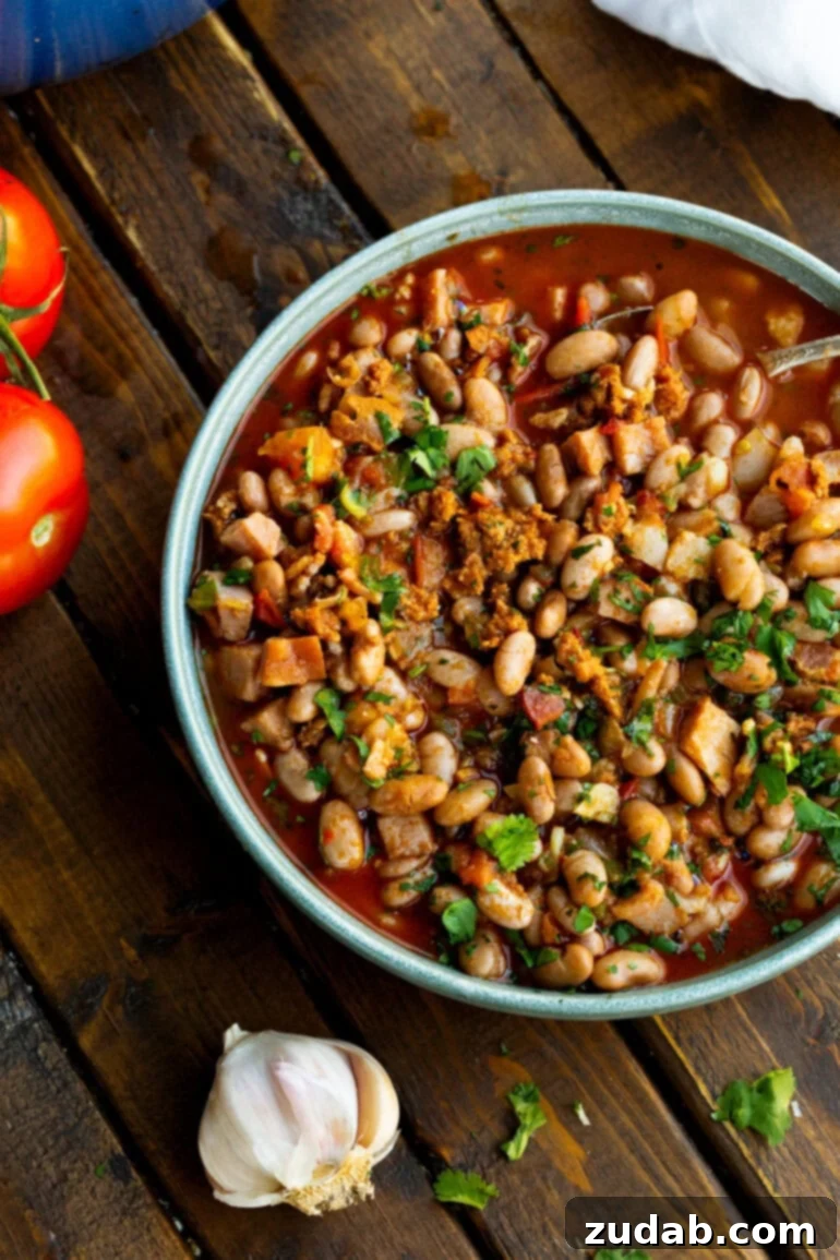 Classic Charro Beans Recipe 11 A rustic bowl of charro beans is beautifully presented on a wooden table, flanked by fresh Roma tomatoes and a head of garlic, highlighting the fresh ingredients and homestyle appeal of the dish.