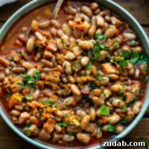 Classic Charro Beans Recipe 13 A bowl of charro beans sitting on a wooden table with with some roma tomatoes and a bulb of garlic sitting next to it