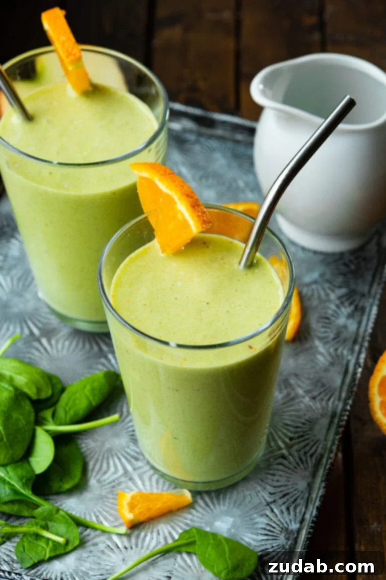 The Ultimate Cold-Busting Green Smoothie 2 A photo of two bright green smoothies, each garnished with a fresh orange wedge and a metal straw, against a light background.