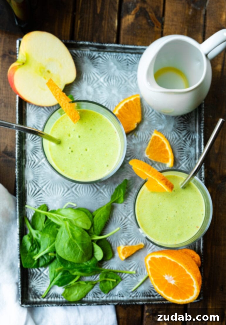 The Ultimate Cold-Busting Green Smoothie 6 An overhead shot of two glasses of bright green smoothie, artfully arranged on a surface surrounded by fresh spinach leaves, orange wedges, and a sliced apple, highlighting the smoothie's vibrant color and natural ingredients.