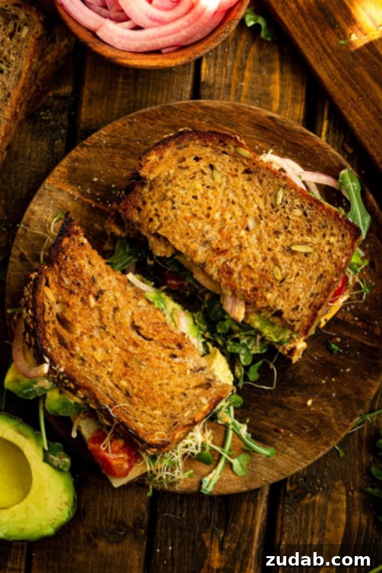 My Signature Avocado Sandwich 3 A top-down view of an avocado sandwich, sliced in half and resting on a rustic wooden plate. The golden-brown toasted bread cradles layers of creamy avocado, vibrant greens, and red elements, hinting at the rich flavors within.