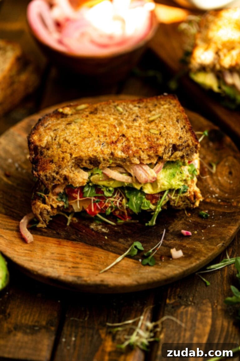 My Signature Avocado Sandwich 6 A perfectly assembled half of an avocado sandwich, showcasing its vibrant layers and golden toasted bread, inviting you to take a delicious bite. It rests on a rustic wooden plate.