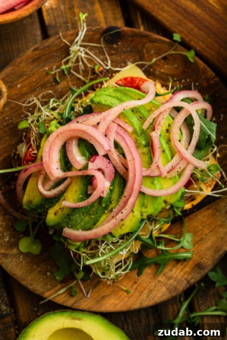 My Signature Avocado Sandwich 7 An appetizing open-faced avocado sandwich, revealing layers of perfectly sliced avocado, bright pink pickled red onions, vibrant microgreens, crunchy bean sprouts, savory cheese, and smoky charred tomatoes on toasted bread.