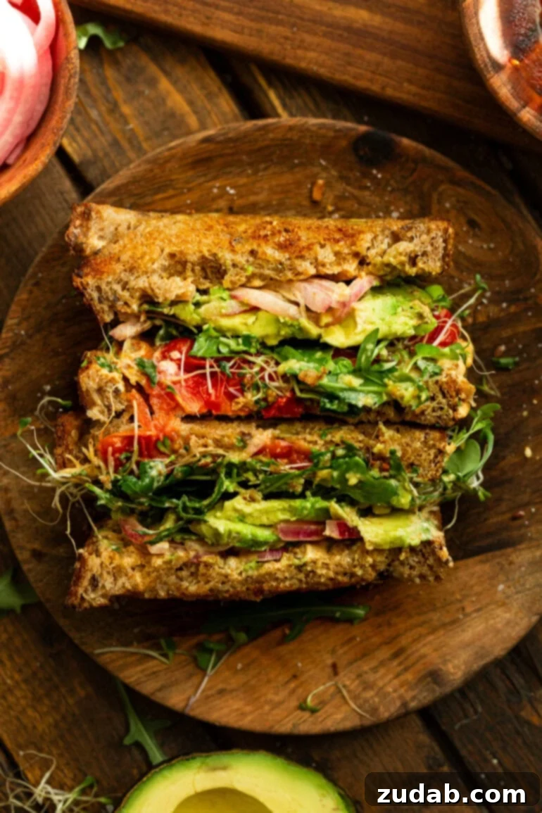 My Signature Avocado Sandwich 9 A sliced avocado sandwich, standing upright to reveal its delicious layers: perfectly toasted bread, creamy avocado, vibrant pickled red onions, fresh sprouts and microgreens, rich Manchego cheese, and smoky charred tomatoes.
