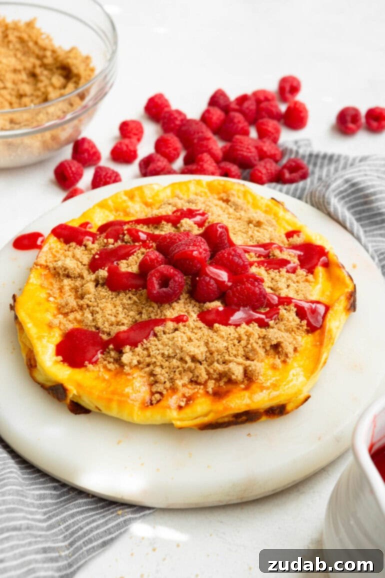 Baked Yogurt Cheesecake 5 A close-up photo of a finished Cheesecake Baked Yogurt, beautifully topped with a generous layer of graham cracker crumble, whole fresh raspberries, and a luscious raspberry sauce drizzle.