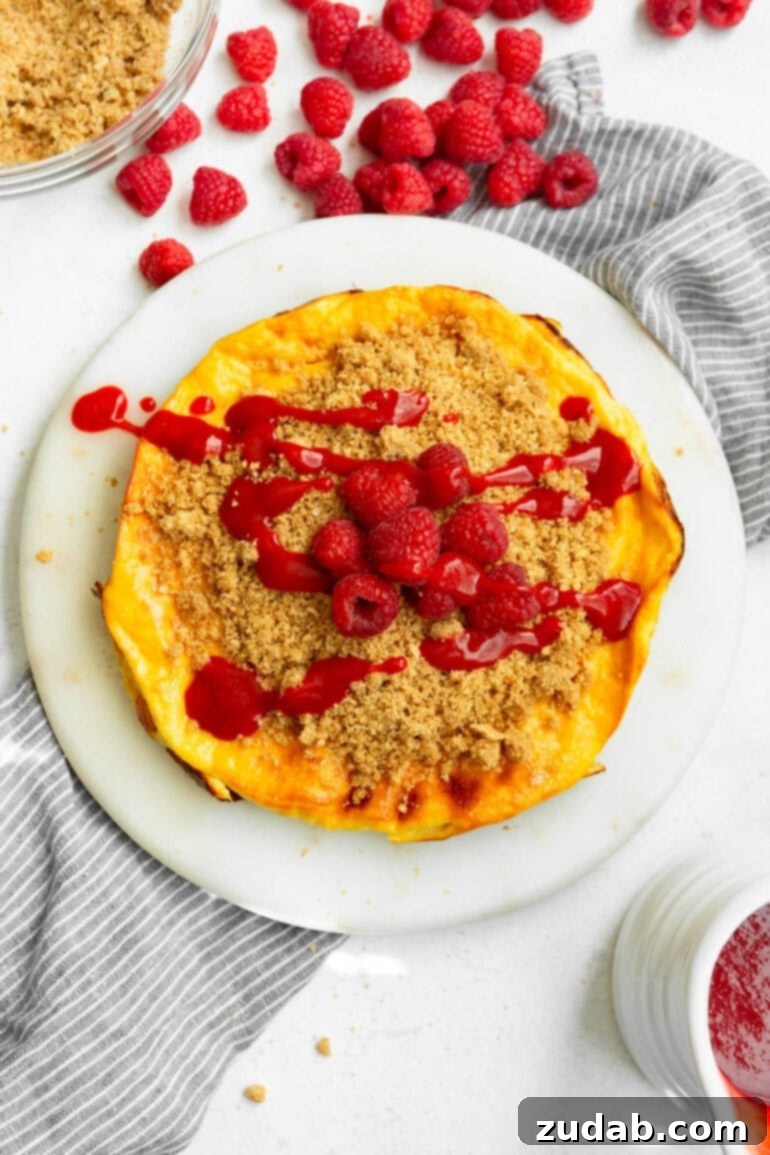 Baked Yogurt Cheesecake 8 A circular baked yogurt, freshly topped with a golden graham cracker crumble, luscious raspberry sauce, and bright whole raspberries, ready to be enjoyed.