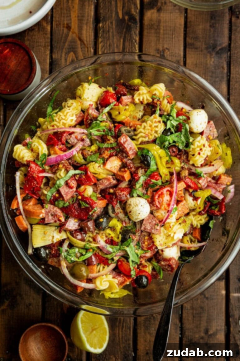 Effortless Antipasto Pasta Salad Recipe 3 A close-up photo of a large glass bowl filled with antipasto pasta salad, featuring cooked pasta, mozzarella pearls, various meats, and vegetables, all generously drizzled with a homemade herb dressing, creating a tantalizing visual of fresh and savory ingredients.