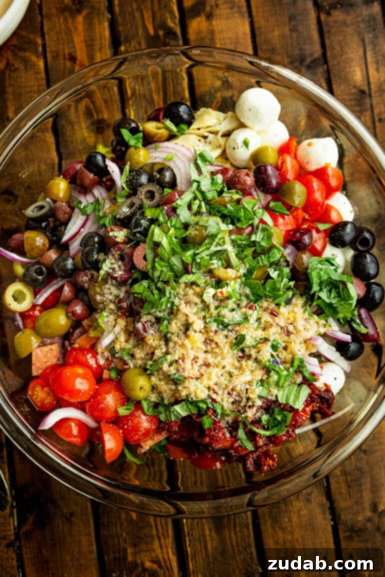 Effortless Antipasto Pasta Salad Recipe 5 A photo showcasing a large glass bowl filled with the unmixed ingredients for antipasto pasta salad, featuring distinct layers of cooked pasta, various meats, cheeses, and vegetables, ready to be tossed together with dressing.