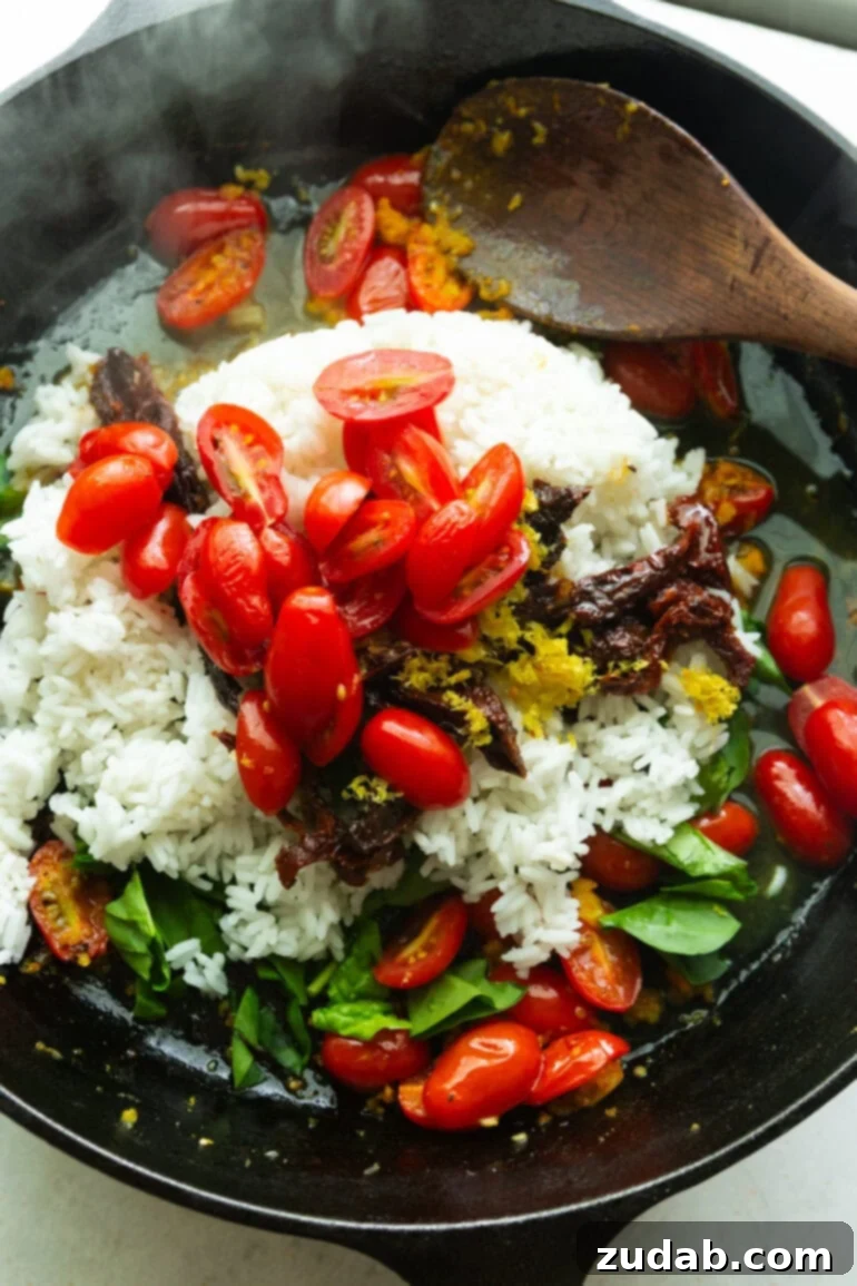 A cast iron skillet is sizzling hot with rice, grape tomatoes, sun-dried tomatoes, aromatic herbs, and olive oil, capturing the essence of Mediterranean cooking.