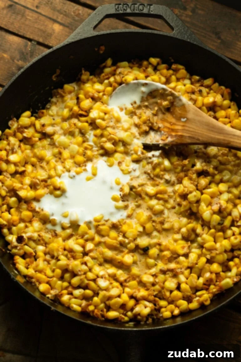 One-Pan Mexican Elote Chicken 7 A cast iron skillet filled with sautéing corn, onions, and spices, with Mexican crema being stirred in by a wooden spoon.