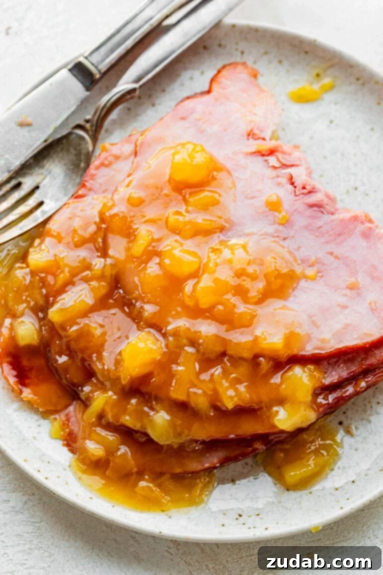 Your New Favorite Slow Cooker Ham 6 a photo of several slices of ham sitting on a plate topped with a pineapple glaze and fork on the side