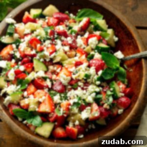 A photo of a large wooden bowl with strawberry cucumber feta salad tossed with fresh herbs
