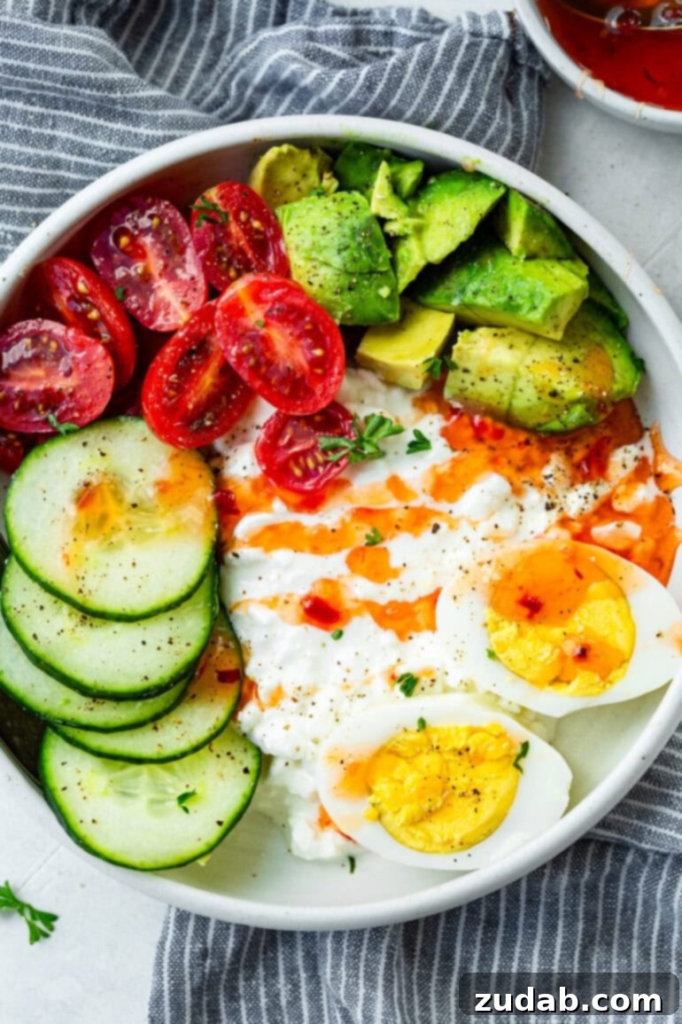 The Ultimate Protein Power Bowl 3 A close-up shot of a cottage cheese bowl topped with neatly arranged hard-boiled egg halves, fresh cucumber slices, avocado chunks, bright cherry tomatoes, and a generous swirl of sweet chili sauce.