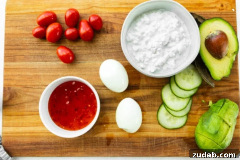 The Ultimate Protein Power Bowl 4 A vibrant flat lay photo showcasing all the fresh ingredients for the high-protein cottage cheese and egg breakfast bowl: a tub of creamy cottage cheese, neatly arranged hard-boiled eggs, bright grape tomatoes, crisp cucumber slices, and a bottle of sweet chili sauce.