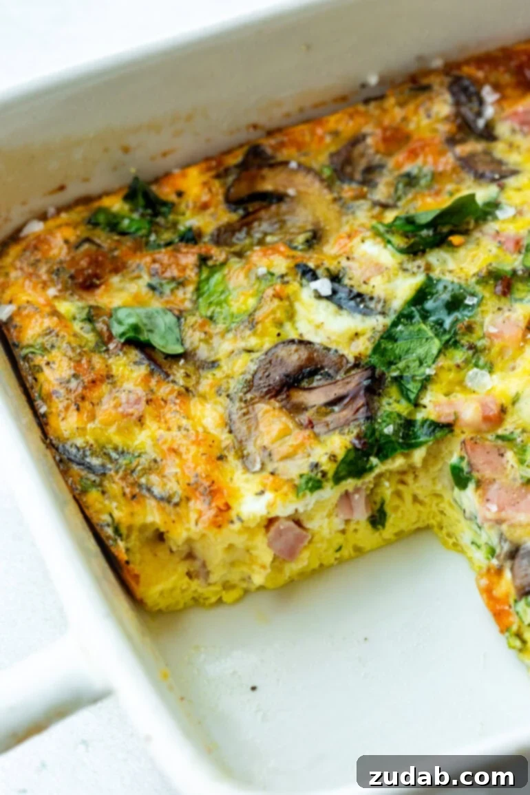 Morning Magic: Ham, Cheese, and Mushroom Egg Bake 2 A baking dish of a baked egg, spinach and sautéed mushroom casserole with a serving missing, showing the cross-section of the casserole.