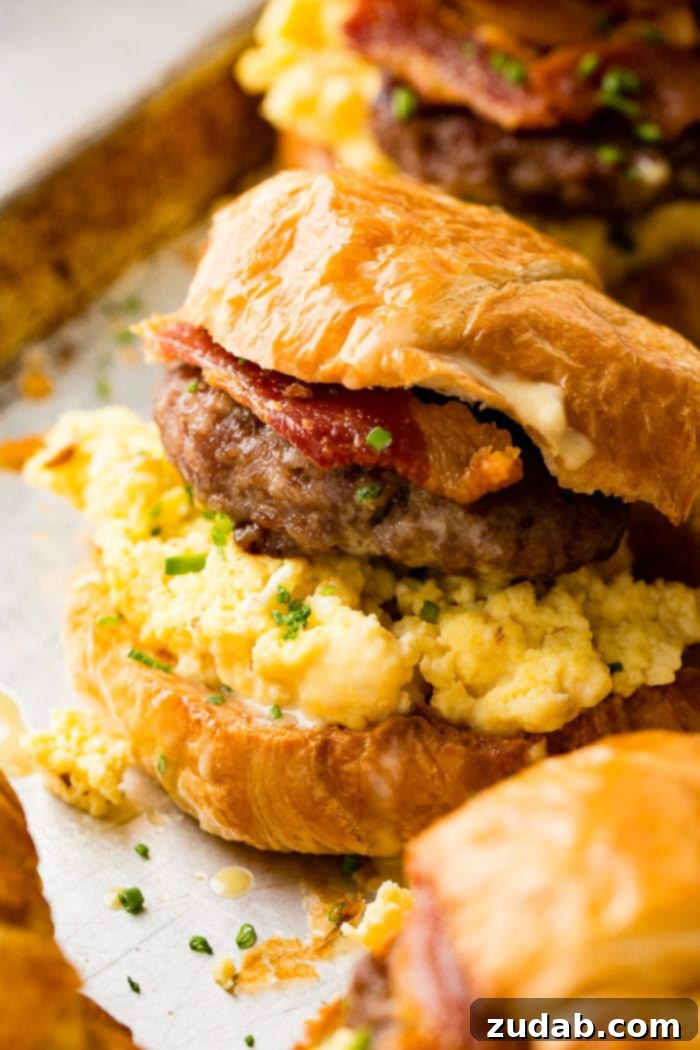 Bacon, Egg, and Cheese Croissant Breakfast Sandwiches 4 A close-up photo of a gourmet breakfast sandwich on a croissant, filled with scrambled eggs, sausage, bacon, and sprinkled with chopped chives.