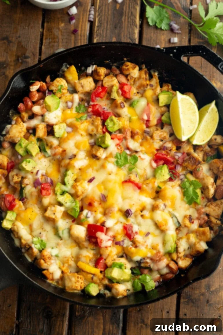 One-Pan Fiesta Chicken & Zucchini Skillet 2 A sizzling cast iron skillet overflowing with golden-brown chicken chunks, melted cheese, fresh avocado slices, sautéed zucchini, and bright lime wedges, ready to be served.