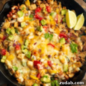 A cast iron skillet full of golden chicken chunks, melted cheese, avocado, zucchini and a couple of lime wedges