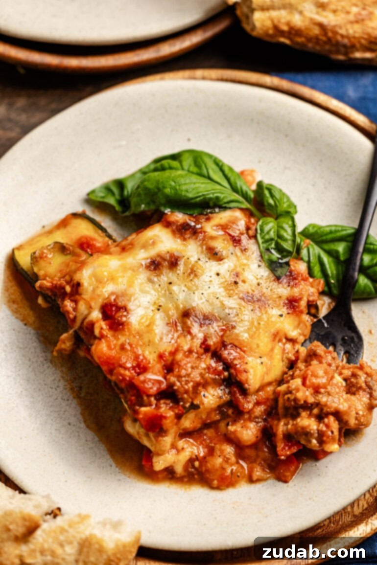 The BEST zucchini lasagna we've ever had! ohsweetbasil.com a plate with a hearty slice of layered zucchini noodles, cheese, and meat sauce with a basil garnish. A fork is digging into the slice of lasagna.