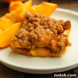 Another view of the peach crumble bar on a white plate with peach slices, highlighting the crisp streusel topping and thick peach layer.