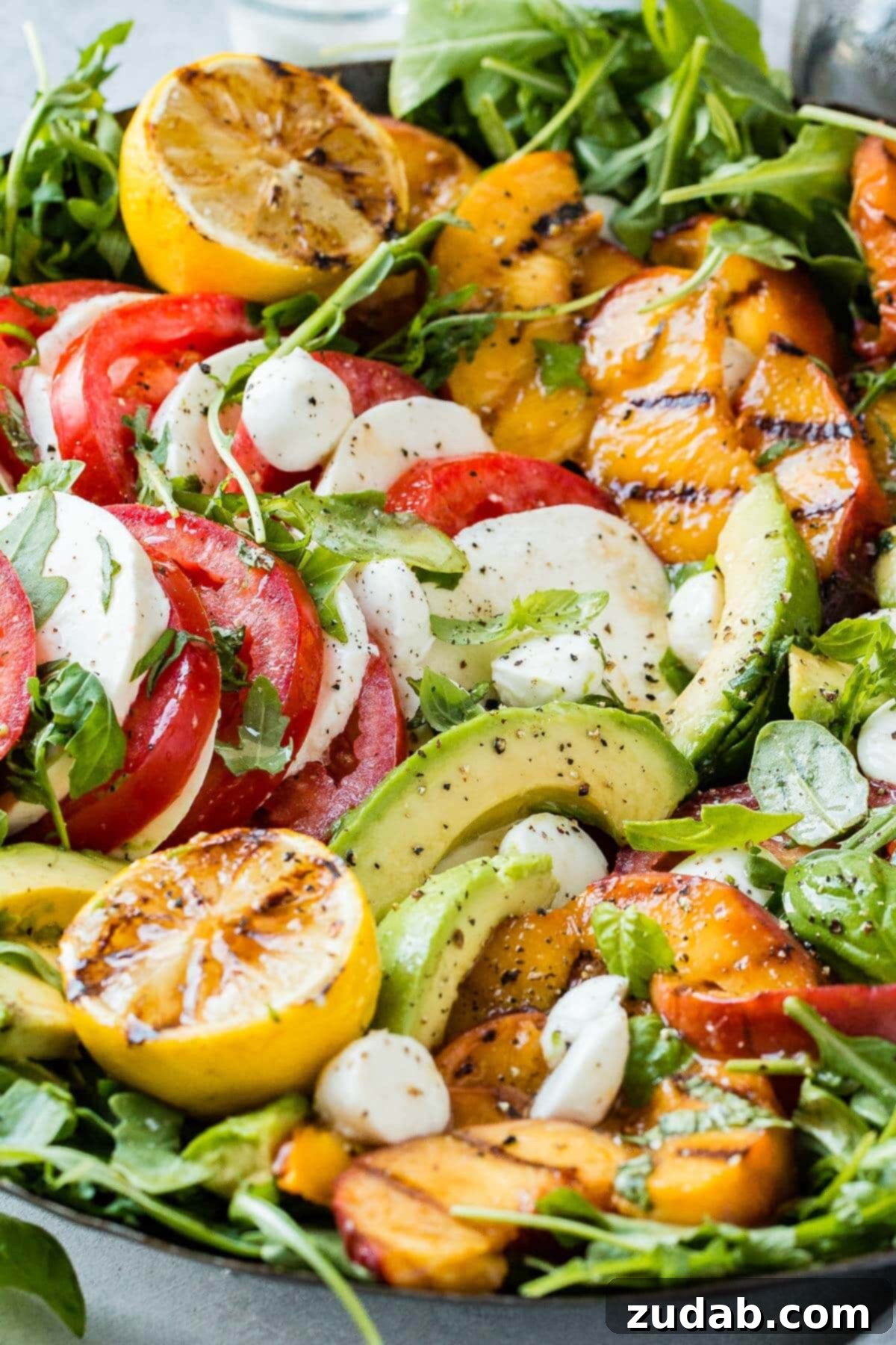 Grilled Peach Caprese 5 Close up of Grilled Peach Caprese Salad filled with slices of avocado, tomato, mozzarella, green leaves, grilled peaches, and two halves of lemons coated with balsamic glaze. This inviting image showcases the freshness and vibrant colors of the summer dish.