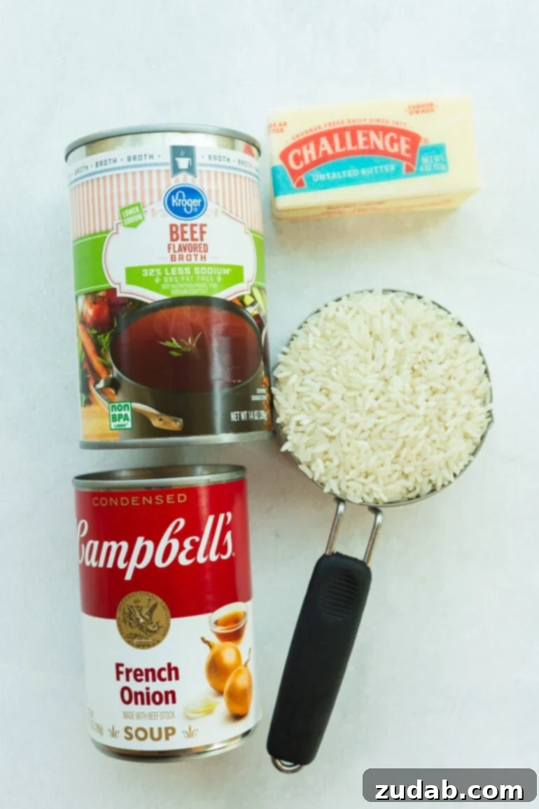 All the ingredients laid out on a counter: long-grain white rice, a stick of unsalted butter, a packet of condensed French onion soup mix, and a can of beef broth. These simple pantry staples are beautifully arranged, highlighting how easily they combine to make a rich, cozy side dish.