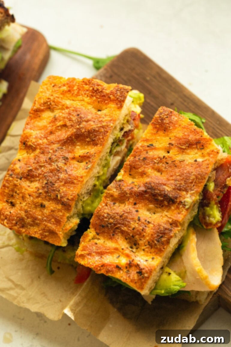 Honey Avocado Turkey Bacon Sandwich 11 A delectable Turkey Bacon Avocado Sandwich on focaccia bread, perfectly cut in half and resting on brown parchment paper, ready to be enjoyed.