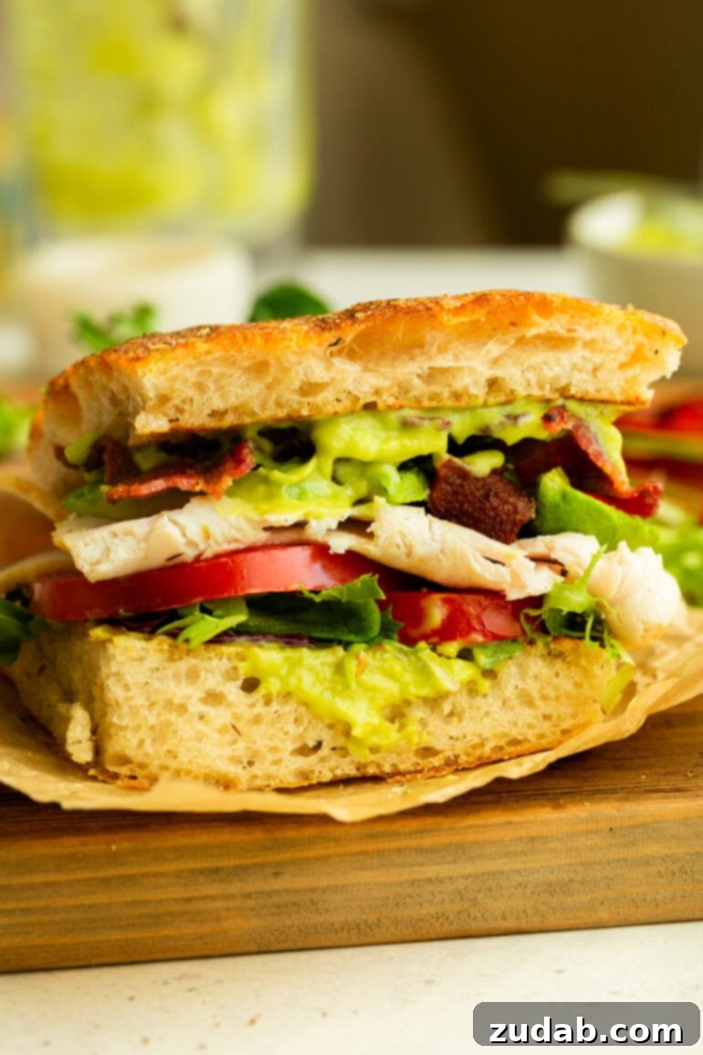Honey Avocado Turkey Bacon Sandwich 5 Half of a delicious Turkey Bacon Avocado Sandwich served on rustic focaccia bread, showcasing its layers of fresh ingredients and creamy honey avocado sauce.