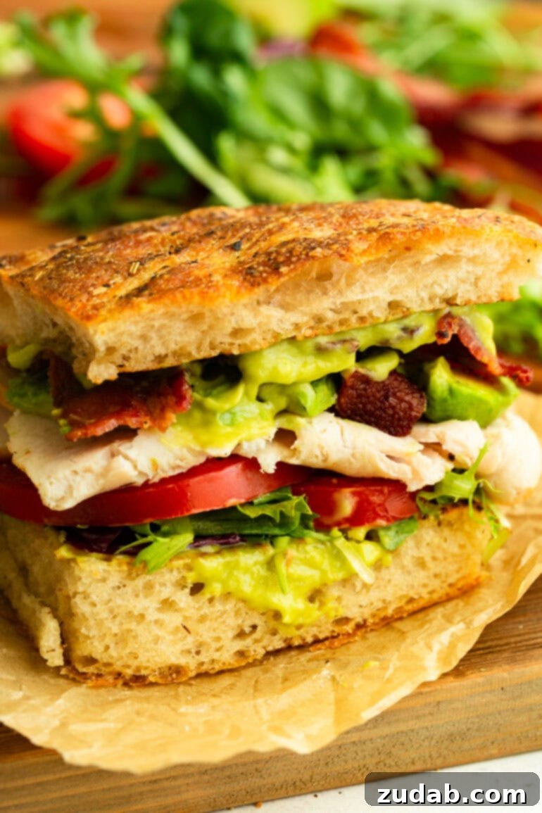 Honey Avocado Turkey Bacon Sandwich 9 A perfectly assembled Turkey Bacon Avocado Sandwich on rustic focaccia bread, showcasing thick slices of turkey, crispy bacon, creamy avocado, and fresh greens, drizzled with honey avocado sauce.