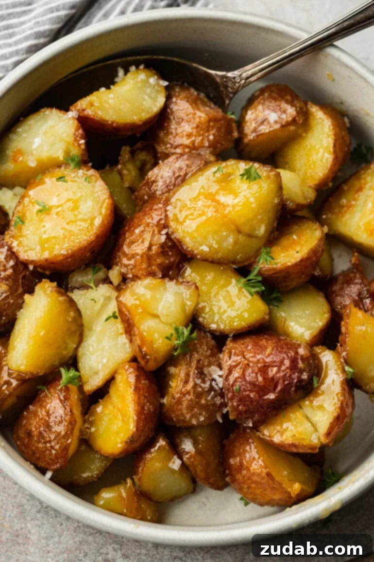 Golden Roasted Red Potatoes 6 A generous serving of golden, crispy roasted red potatoes is presented in a clean white bowl, accompanied by a silver spoon. Each potato glistens with a honey butter Dijon glaze, delicately sprinkled with flaky sea salt.