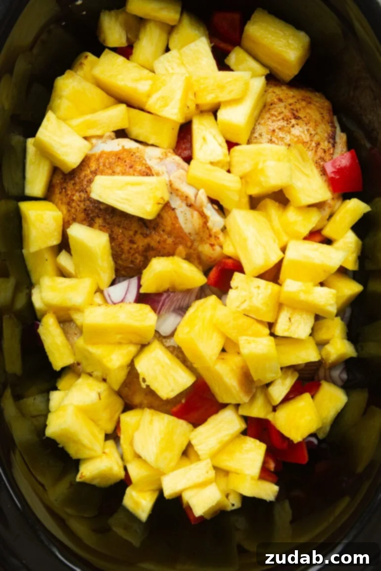 Sweet and Tangy Slow Cooker Pineapple Chicken 3 A black crockpot filled with pan-seared chicken thighs, vibrant pineapple chunks, chopped red bell peppers, and purple onion, simmering in a rich, savory sauce
