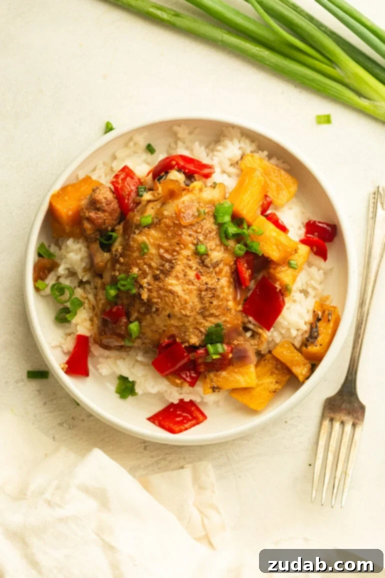 Sweet and Tangy Slow Cooker Pineapple Chicken 7 A white plate featuring a generous serving of white rice, topped with a succulent golden pineapple chicken thigh, surrounded by saucy pineapple, red bell pepper, and onions, all garnished with fresh chopped green onions