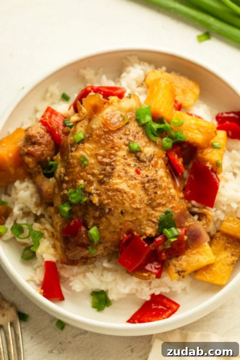 Sweet and Tangy Slow Cooker Pineapple Chicken 8 A dinner plate featuring a serving of white rice with a succulent BBQ teriyaki chicken thigh and generous portions of pineapple, bell peppers, and onions, all generously coated in sauce