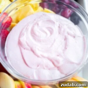 A small glass full of light pink thick and creamy strawberry fluff fruit dip, surrounded by cut fruit for dipping.