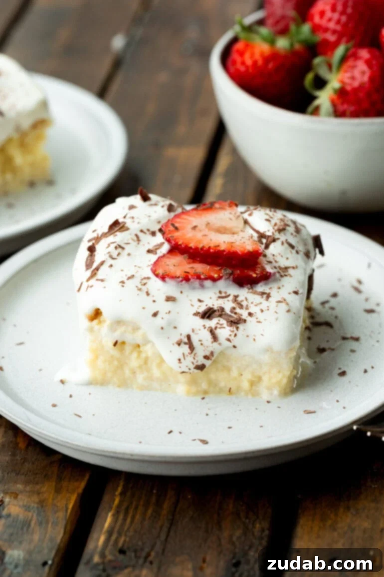 Mexican Tres Leches Cake Recipe 2 a photo of a slice of tres leches cake on a small dessert plate topped with two slices of strawberry and shaved chocolate