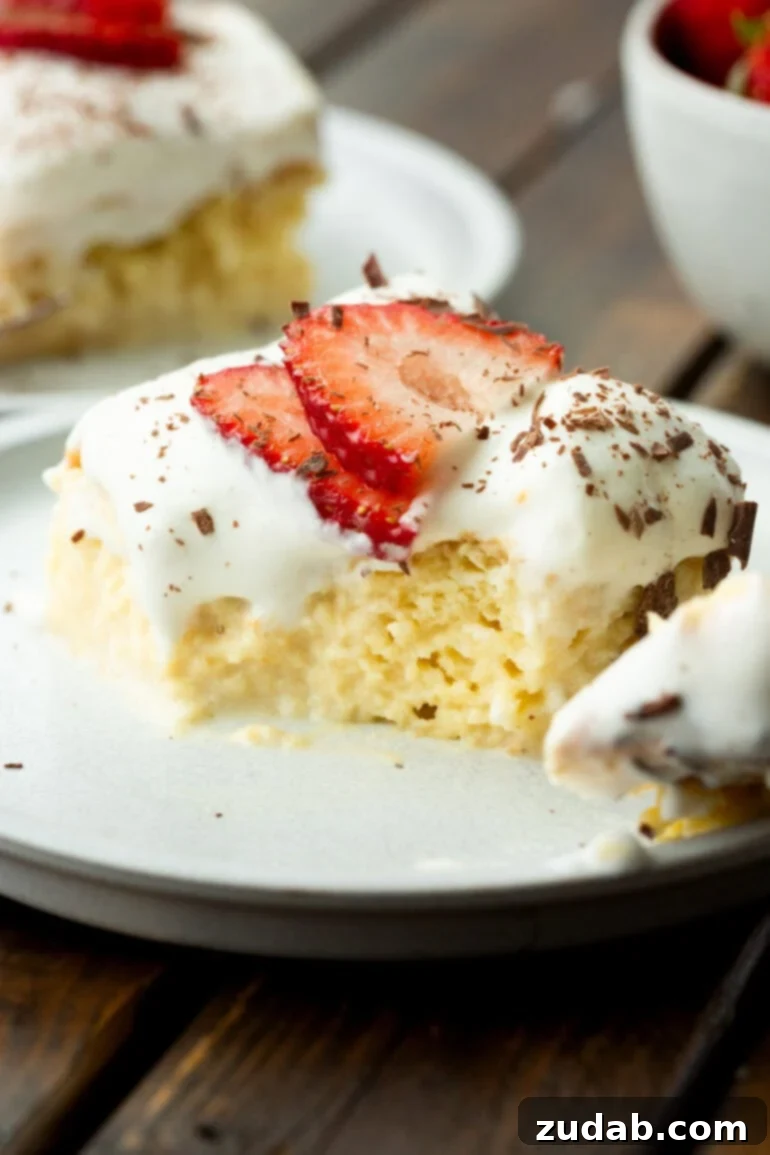 Mexican Tres Leches Cake Recipe 5 a photo of a pieces of tres leches cake with a bite removed so you can see the inside of the moist cake topped with whipped cream, sliced strawberries and shaved chocolate