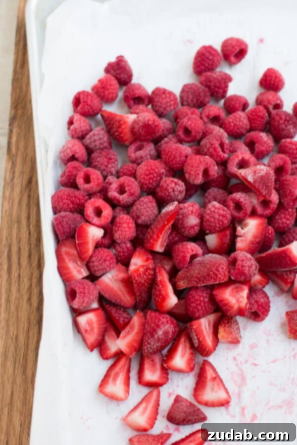 how to freeze fruit and make better and cheaper smoothies ohsweetbasil.com a photo of several whole raspberries and quartered strawberries that are frozen and sitting on a baking sheet lined with parchment paper