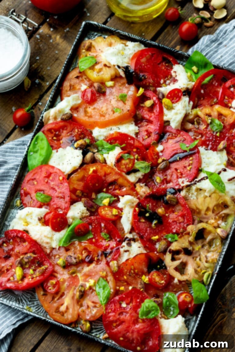 A beautifully arranged Caprese salad spread over a serving tray, featuring thick slices of red, orange, and green heirloom tomatoes, fresh mozzarella medallions, bright green basil leaves, a generous sprinkle of toasted pistachios, and a glistening balsamic drizzle, seasoned lightly with salt and pepper.