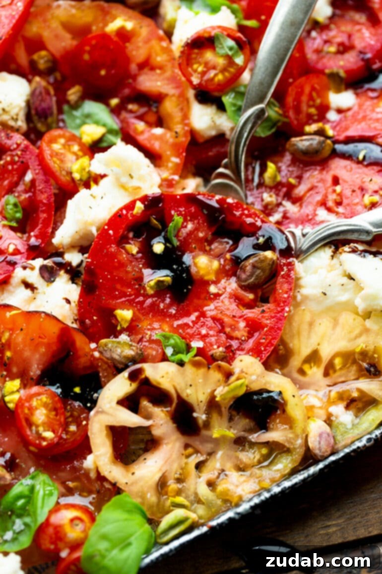 Close-up photo of vibrant sliced heirloom tomatoes in various colors, alongside chunks of fresh mozzarella, interspersed with fresh basil leaves, all glistening under a balsamic glaze and topped with chopped pistachios.