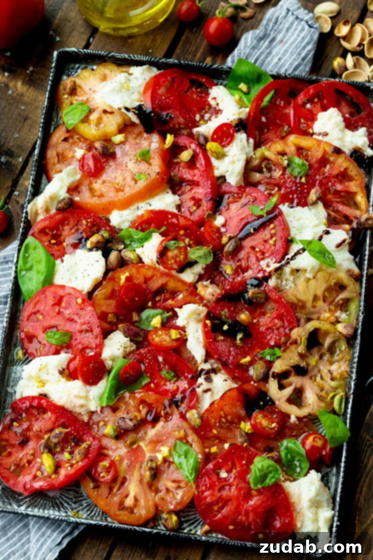 A large serving tray generously filled with vibrant sliced tomatoes and substantial chunks of fresh mozzarella, elegantly topped with fresh basil leaves, a rich balsamic drizzle, and scattered toasted pistachios, creating a visually appealing and delicious Caprese salad.