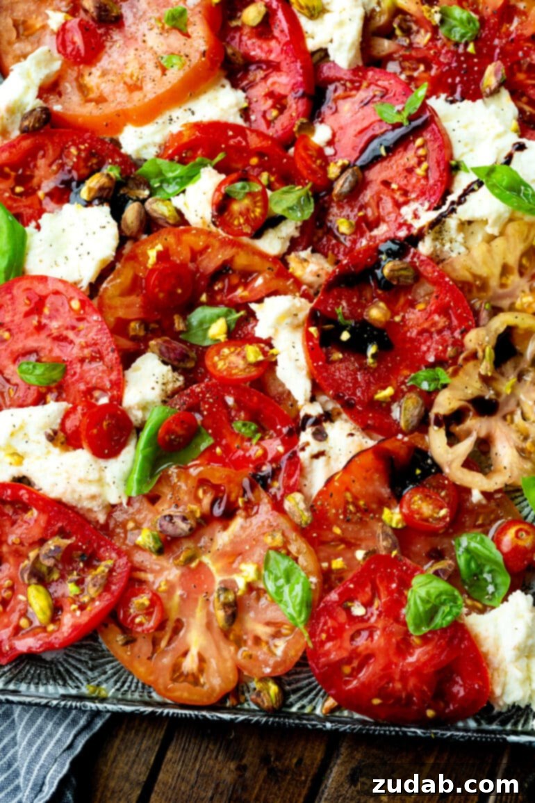 A large serving tray piled high with Caprese salad, featuring generous slices of tomatoes and fresh mozzarella, adorned with fresh basil leaves, a generous scattering of toasted pistachios, and a rich balsamic drizzle, ready to be enjoyed.