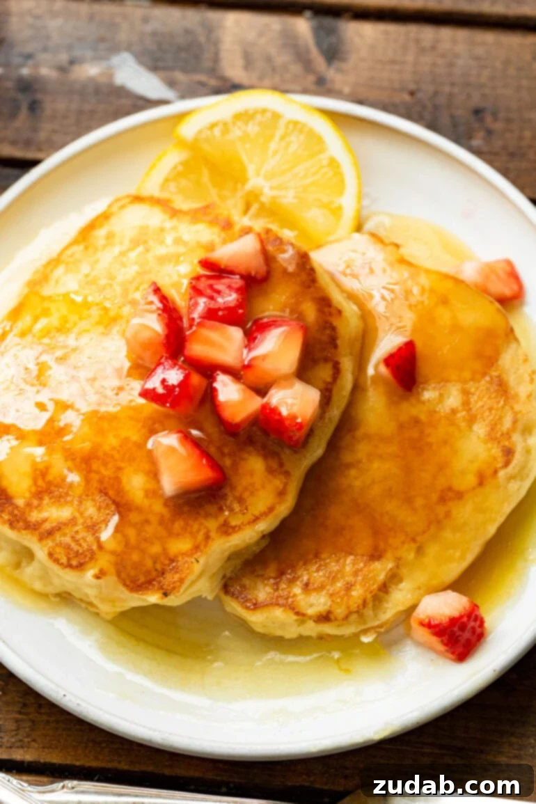 Melt-Away Buttermilk Pancakes 2 A vibrant photo showcasing several perfectly fluffy golden buttermilk pancakes elegantly stacked on a pristine white plate. The pancakes are generously adorned with fresh, diced strawberries and exquisitely drizzled with warm, homemade buttermilk syrup, highlighting their irresistible melt-in-your-mouth quality and inviting presentation.