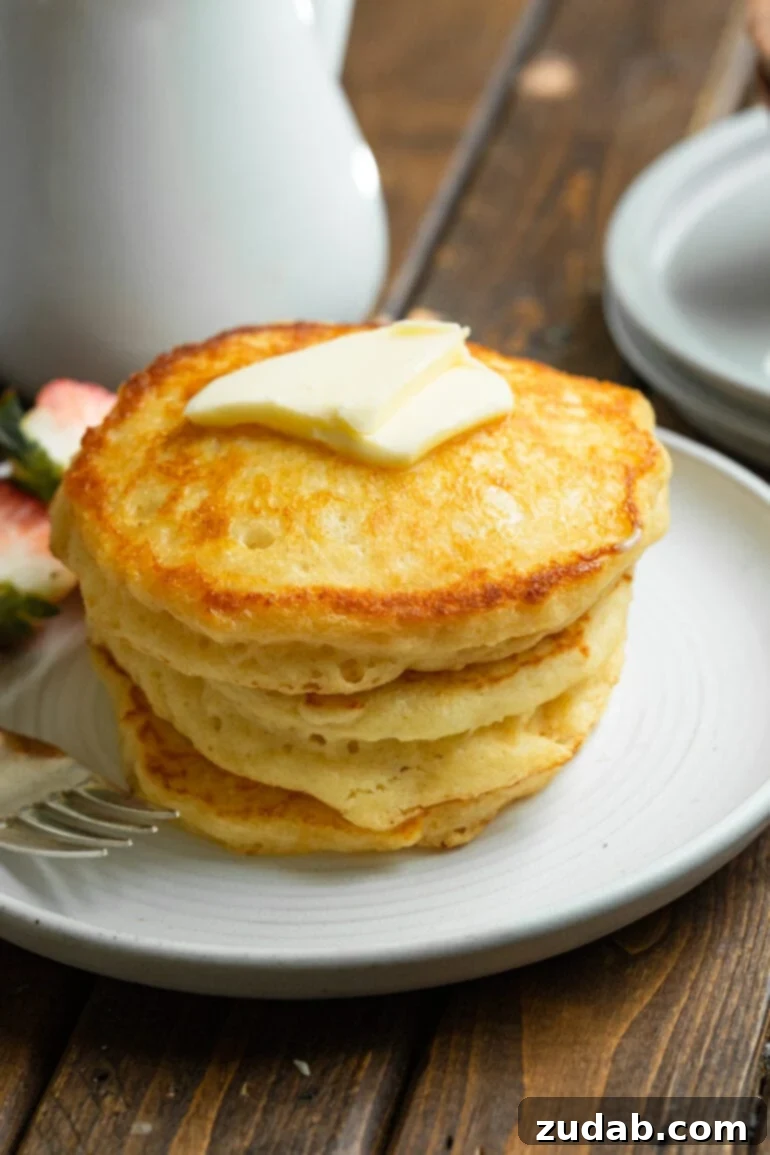 Melt-Away Buttermilk Pancakes 5 A captivating photo of a perfectly balanced stack of four golden, gloriously fluffy buttermilk pancakes, resting elegantly on a pristine white plate, luxuriously crowned with a small pat of melting butter. This image embodies the sheer comfort and simple elegance of a perfect Sunday morning breakfast, featuring our irresistible melt-in-your-mouth buttermilk pancakes, promising a moment of pure culinary bliss.