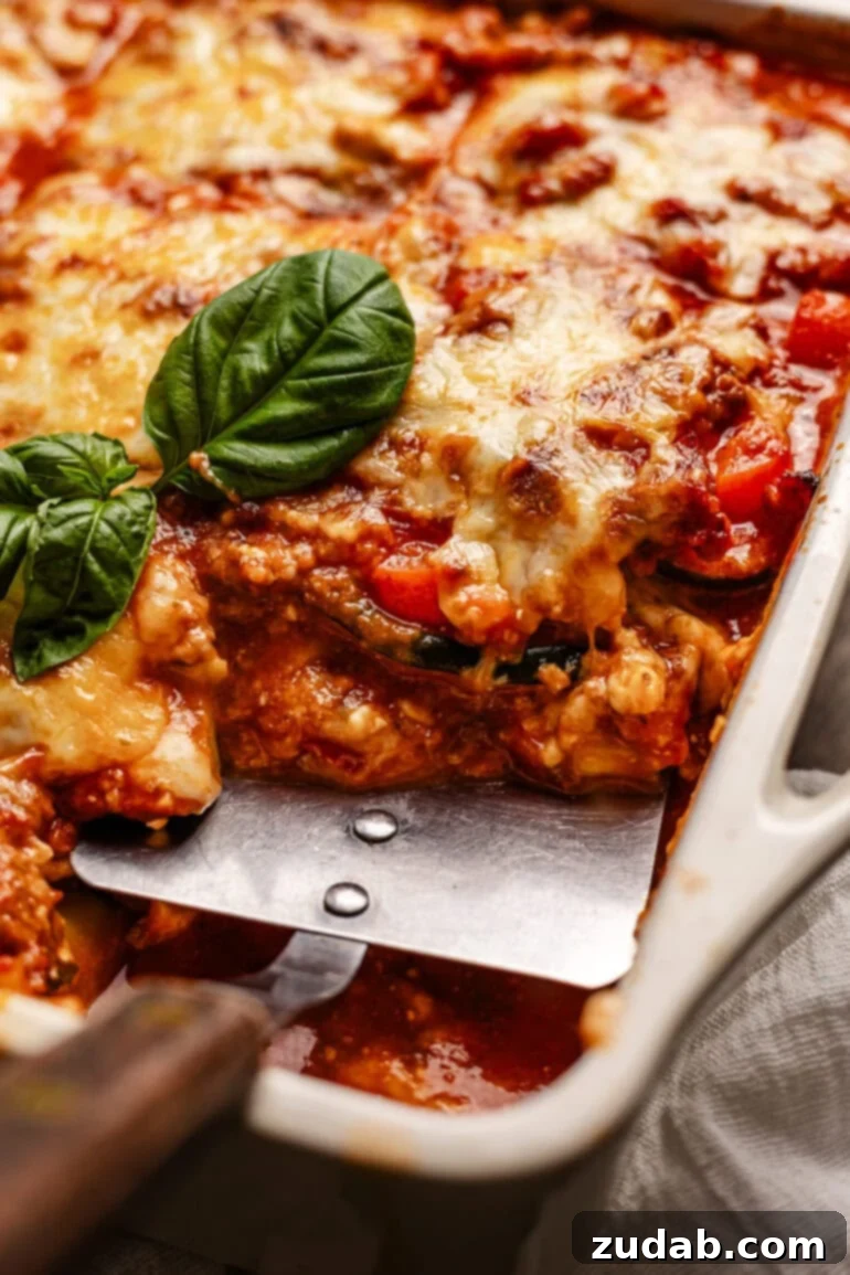 The BEST zucchini lasagna we've ever had! ohsweetbasil.com A metal spatula is digging into a full baking dish of layered veggies, meat sauce, and cheese, showcasing the rich texture of Zucchini Lasagna.