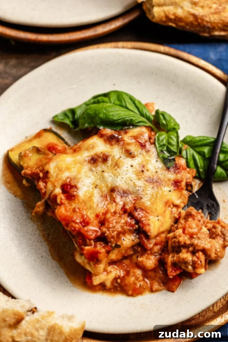 The BEST zucchini lasagna we've ever had! ohsweetbasil.com A plate with a hearty slice of layered zucchini noodles, cheese, and meat sauce with a basil garnish. A fork is digging into the slice of lasagna, inviting you to taste.