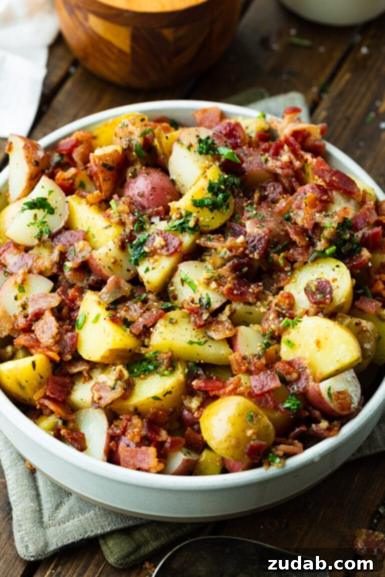 A close-up photo of a large white bowl filled with German potato salad, showcasing cubed potatoes, generous bacon bits, and freshly chopped parsley.