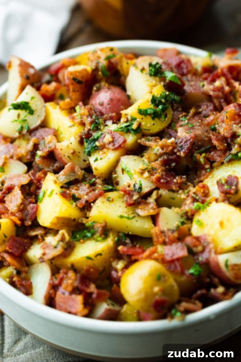 A close-up photo of a bowl of warm German potato salad, generously loaded with crispy bacon crumbles, minced garlic, and fresh green parsley.