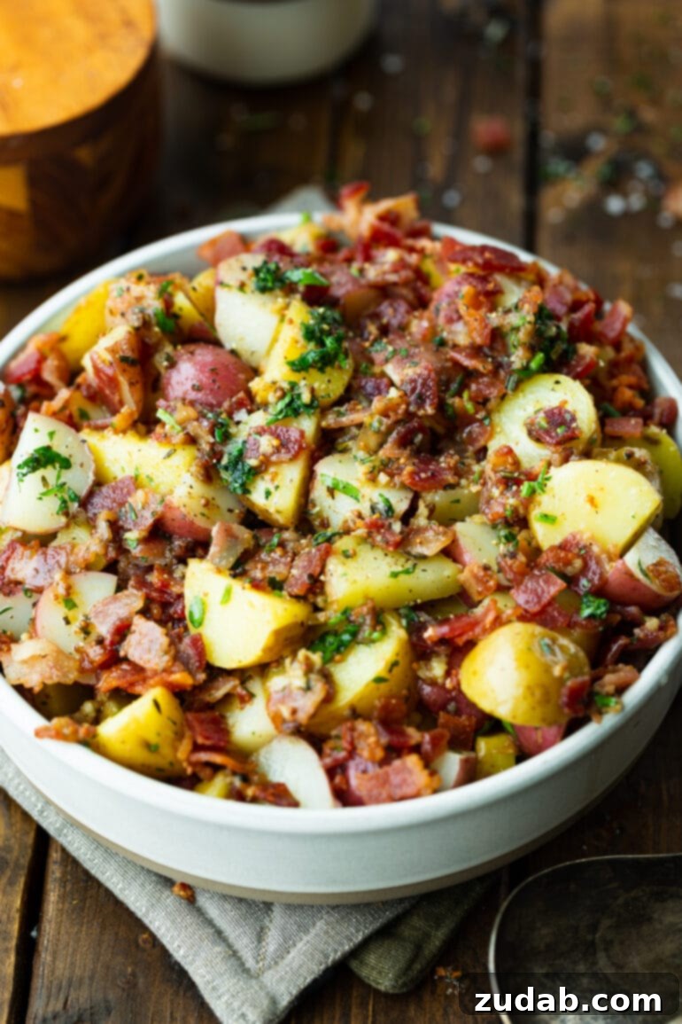 A photo of German potato salad, featuring cooked potatoes, crunchy bacon crumbles, garlic, and fresh parsley, presented in a large white serving bowl.