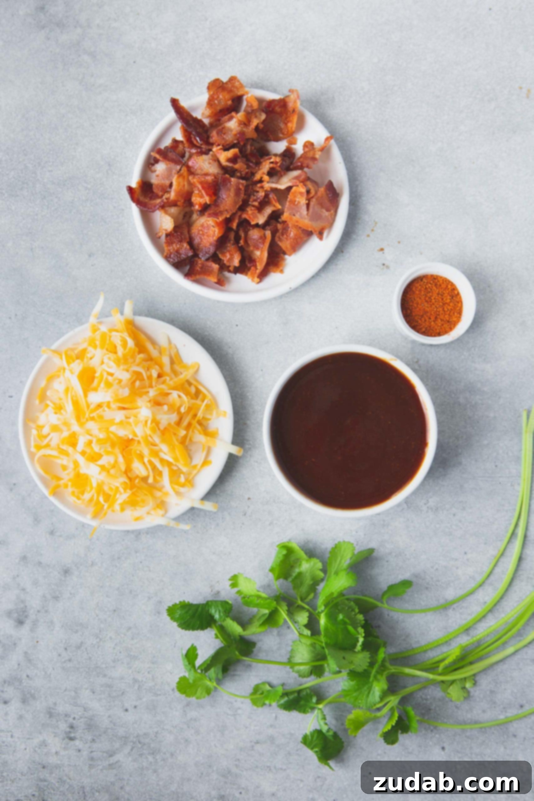 Various ingredients for Cheesy Bacon BBQ Chicken displayed in individual bowls, including grated cheese, a rich BBQ sauce, crispy bacon, and a selection of spices.