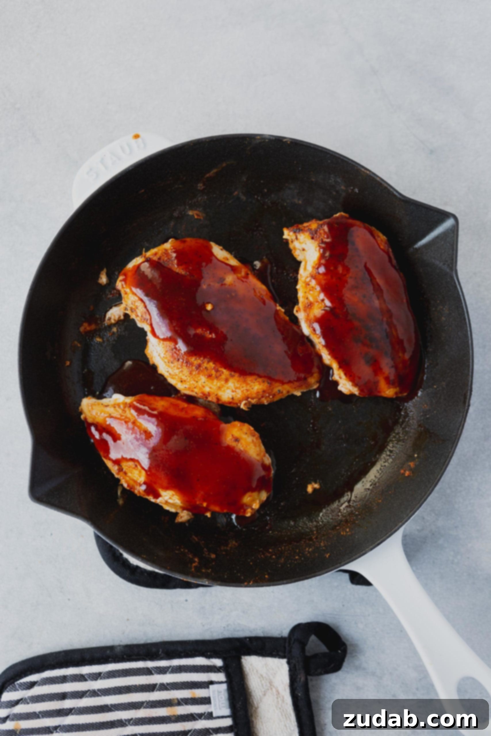 Three perfectly seared chicken breasts arranged in a skillet, already coated with a generous layer of rich BBQ sauce.